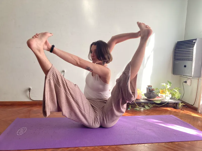 Eugenia practicando en su casa sobre el mat, sentada en equilibrio tomando sus pies con las piernas extendidas hacia arriba.